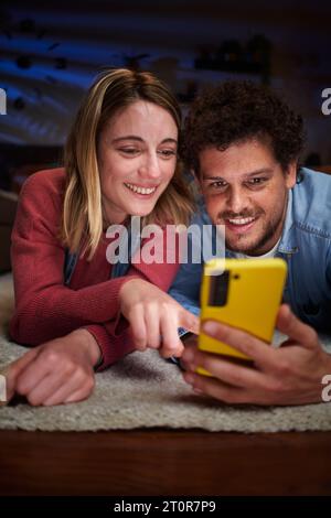 Vertikales glückliches kaukasisches Paar mit Telefon lächelnd auf Teppich im Wohnzimmer zu Hause liegend. Stockfoto