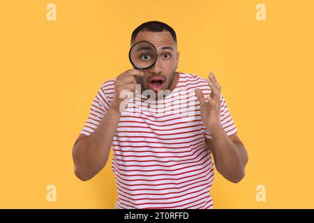 Schockierter Mann, der durch die Lupe auf orangem Hintergrund schaut Stockfoto