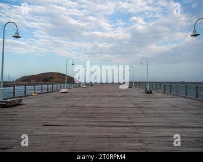 Direkt den Coffs Harbour Jetty Pier hinunter, gesäumt von Straßenlaternen und Sitzplätzen auf jeder Seite, MuttonBird Island im Hintergrund, Australien Stockfoto