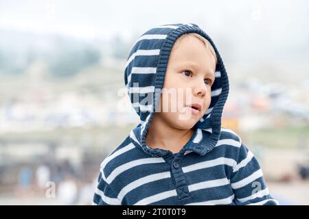 Der verspielte kleine Junge trägt einen Hoodie, der im Freien ein Gesicht auf unscharfem Hintergrund zaubert. Lustiges Kind, das Kopf auf Schulter geht, geht im Bergpark. Glückliche Kindheit Stockfoto