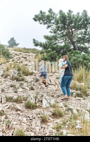 Mutter trägt das Baby, das mit einem kleinen Sohn im wilden Naturpark auf den felsigen Hügel geht. Familie von Eltern und Kindern spazieren im Hochland. Frau mit Kindern Stockfoto