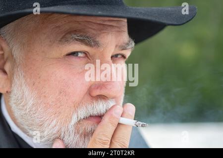 Nahaufnahme Porträt eines seriösen, stylischen Hipster-Großvaters im Ledermantel, der draußen raucht. Älterer Großvater, Großvater Rentner, Rentner-Konzept. Stockfoto