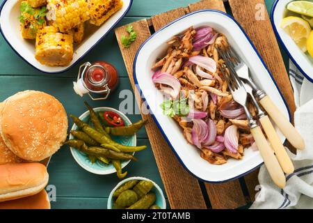 Gezapftes bbq-Hühnchen mit gebackenen Zwiebeln auf Servierplatte, bbq-Mais, Gurken, Chilischoten und Brötchen für Hot Dogs und Burger, Tomatensauce. Traditionell Stockfoto