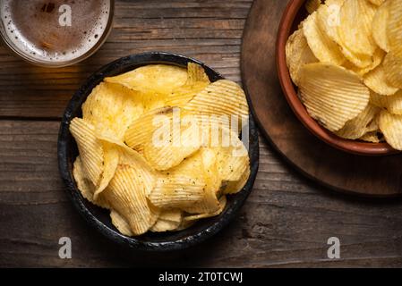 Knusprige Kartoffelchips in einer Schüssel und leichtes Bier in einem Glas auf hölzernem Hintergrund Stockfoto