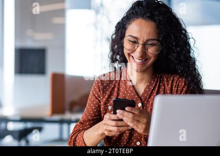 Schöne Geschäftsfrau am Arbeitsplatz, erfolgreiche und fröhliche lateinamerikanische Frau, die per App SMS auf dem Smartphone eingibt, Nahaufnahme weibliche Arbeiterin, die zufrieden lächelt. Stockfoto
