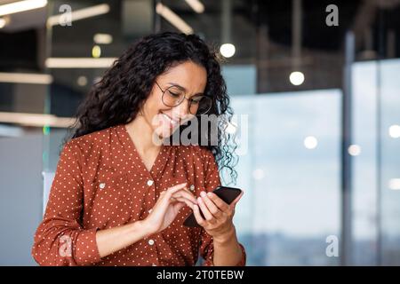 Schöne Geschäftsfrau am Arbeitsplatz, erfolgreiche und fröhliche lateinamerikanische Frau, die per App SMS auf dem Smartphone eingibt, Nahaufnahme weibliche Arbeiterin, die zufrieden lächelt. Stockfoto