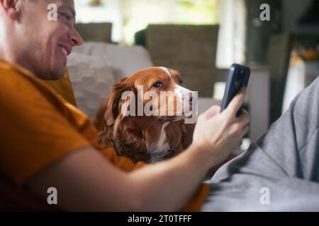 Mann sitzt auf Sofa mit Hund. Haustierbesitzer macht Selfie über das Handy mit seinem Nova Scotia Duck Tolling Retriever zu Hause. Stockfoto