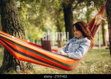 Schöne junge Frau, die auf Laptop im Internet surft, während sie in der Hängematte liegt, malerischer tropischer Park im Hintergrund. Stockfoto