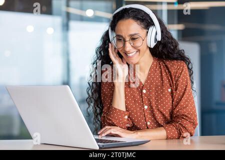 Junge schöne Frau in Kopfhörern, die mit Laptop im Büro am Arbeitsplatz arbeitet, freudige und zufriedene Geschäftsfrau, die Online-Webinar, Lehrvideos, Fernlernen ansieht. Stockfoto