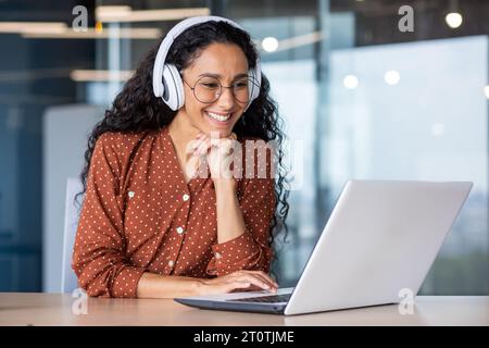 Junge schöne Frau in Kopfhörern, die mit Laptop im Büro am Arbeitsplatz arbeitet, freudige und zufriedene Geschäftsfrau, die Online-Webinar, Lehrvideos, Fernlernen ansieht. Stockfoto