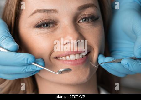 Schöne und schöne Frau beim Zahnarzttermin. Nahaufnahme des Gesichts, das in die Kamera blickt und lächelt. Zahnarzthände mit Instrumenten in Patientennähe Stockfoto