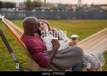 Liebevolles Paar umarmt sich, während es draußen in einer Hängematte sitzt, in eine warme Decke gehüllt. Schwarzer Mann und kaukasische Frau, die tief verliebt sind, genießen gemütliche M Stockfoto