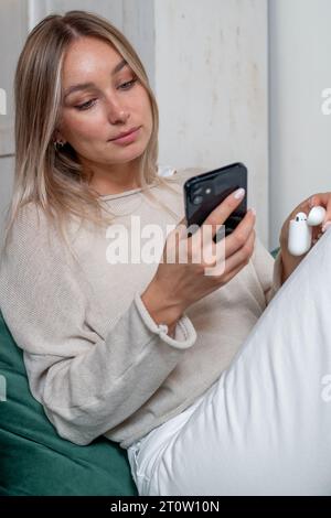 Weiße Frau mit blonden Haaren, die ihr Telefon benutzt, während sie zu Hause auf dem Sofa sitzt Stockfoto