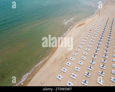 Am Meer erstreckt sich ein Sandstrand, der mit leeren Liegestühlen geschmückt ist. Aus der Luftperspektive entfaltet sich die ruhige Szene mit dem azurblauen Meer Stockfoto