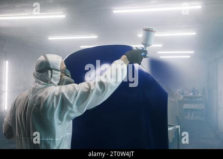 Der Arbeiter lackiert Karosserieteile in der Werkstattkammer mit Airbrush. Mann mit Spritzpistole beim automatischen Lackieren. Stockfoto