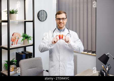 Arzt in weißer Jacke und Brille hält ein rotes Herz in der Krankenhauspraxis. Medizinisches Versorgungskonzept und Ärztedienstkonzept. Stockfoto