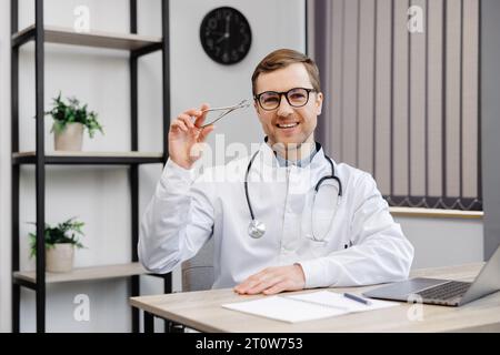 Junger attraktiver Hals-Nasen-Ohren-Arzt, der an seinem Arbeitsplatz in der Praxis sitzt und einen Nasenspiegel in der Hand hält. Stockfoto