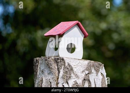 Weißes altes Vogelhaus mit gemustertem rotem Dach steht auf einem Baumstumpf Stockfoto
