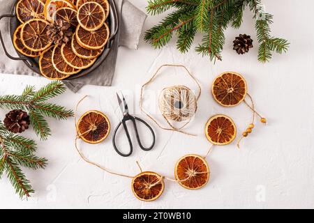 Blick von oben auf weißen strukturierten Hintergrund mit hausgemachter Girlande aus getrockneten Orangen, Fichtenzweigen, Orangenscheiben und handwerklicher Schere. Flache Lagen. Dekoration Stockfoto