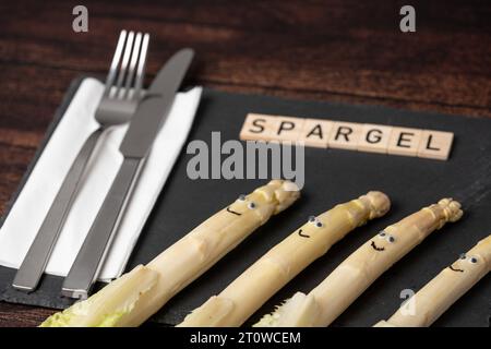 Augsburg, Bayern, Deutschland - 16. April 2023: Spargel mit lächelnden Gesichtern auf einem Teller aus Schiefer mit Messer und Gabel und Holzwürfeln mit Schrift Spargel *** Spargel mit lächelnden Gesichtern auf einem Teller aus Schiefer mit Messer und Gabel und Holzwürfeln mit Schrift Spargel Stockfoto