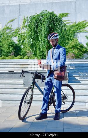 Junger bärtiger Geschäftsmann mit Fahrrad mit Kaffeetasse und digitalem Tablet. Business- und Alternativverkehrskonzept Stockfoto