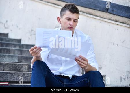 Junger Geschäftsmann, der auf einer Treppe sitzt und Finanzdaten und Diagramme auf Dokumenten analysiert. Handels- und Erfolgskonzept. Stockfoto