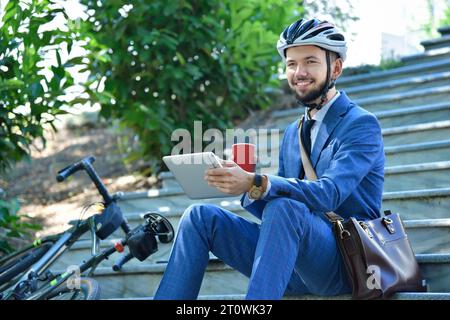 Glücklicher bärtiger Geschäftsmann mit Fahrrad, der auf einer Treppe mit Kaffeetasse und digitalem Tablet sitzt. Business- und Alternativverkehrskonzept Stockfoto