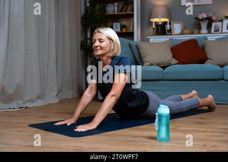 Seniorin, die in Lotusposition sitzt. Aktive reife Frau, die zu Hause im Wohnzimmer Dehnübungen macht. Spannarm für Damen Stockfoto