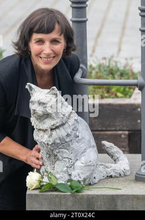 Memmingen, Deutschland. Oktober 2023. Die Künstlerin Cornelia Brader steht neben der Bronzeskulptur der Katze Chicco, die sie gemacht hat. Die burmesische Katze war in der schwäbischen Stadt bekannt und sehr beliebt. Anfang 2023 starb Chicco. Als Ergebnis wurden Spenden für das Denkmal gesammelt, das ihm am Stadtstrom gedenken soll. Quelle: Stefan Puchner/dpa/Alamy Live News Stockfoto