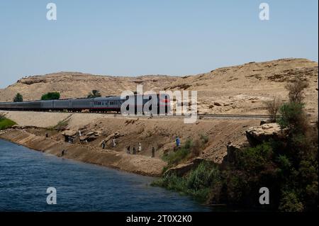 Rote, weiße und schwarze Züge fahren entlang des Nils, während die Einheimischen fleißig auf den Hängen arbeiten, die zum Nil führen. Stockfoto