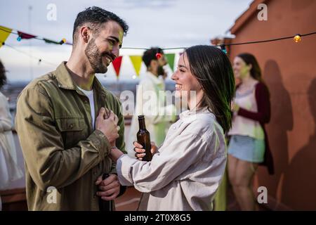 Schönes Paar, das Hände hält und auf einer Dachparty lächelt. Junger und sorgloser Mann und Frau, die sich abends auf einer Partyterrasse anschauen. Stockfoto