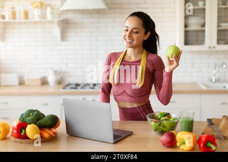 Lächelnde Fitness-Ernährungsberaterin mit Laptop und Apfel in der Küche Stockfoto