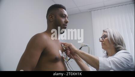 Die Kardiologin, Doktor, setzt holter-Sensoren auf Patienten mit Herz-Kreislauf-Erkrankungen. Afroamerikaner während der funktionellen Rehabilitation im Kardiologiesaal. Elektrokardiographie im modernen Krankenhaus Stockfoto