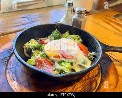 Eine Nahaufnahme mit einer herzhaften Mahlzeit in einer gusseisernen Pfanne auf einem rustikalen Holztisch, mit Spiegeleiern, Brokkoli und Tomaten, die das Wesen eines verkörpern Stockfoto