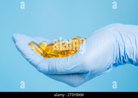 Arzt, Apotheker oder Wissenschaftler Hand in Handschuh mit Omega-3-Kapseln auf blauem Hintergrund. Fischöl-Vitamine und Nahrungsergänzungsmittel in Therapie oder Wissenschaft Stockfoto