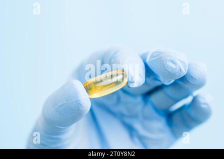 Arzt, Apotheker oder Wissenschaftler Hand in blauen Handschuhen mit Omega-3-Kapsel. Vitaminpille oder -Ergänzung auf blauem Hintergrund. Gesundheitswesen und Wissenschaft Stockfoto