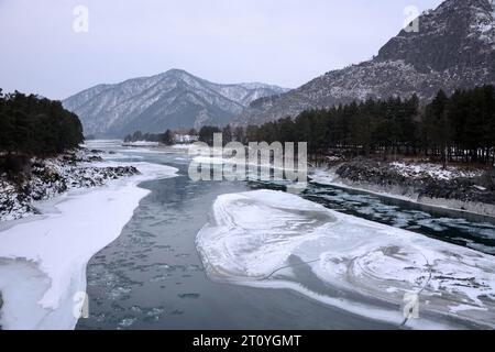 Das geschmolzene Bett eines kleinen Flusses, der in einer Steinhülle aus den Bergen fließt, auf hohen, schneebedeckten Bergen an einem bewölkten Wintermorgen. Fluss Katun, A Stockfoto