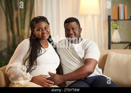 Porträt des lächelnden Ehemanns und seiner schwangeren Frau, die zu Hause auf der Couch sitzen Stockfoto