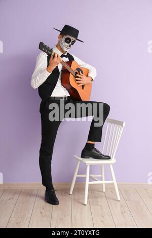 Junger Mann mit bemaltem Schädel, der Gitarre in der Nähe der Fliederwand spielt. Mexikanischer Tag der Toten (El Dia de Muertos) Stockfoto