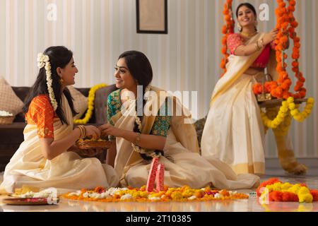 Zwei Frauen dekorieren den Boden mit Blumen auf Onam, während andere Frauen auf einer Schaukel sitzen Stockfoto