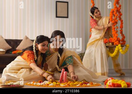 Zwei Frauen dekorieren den Boden mit Blumen auf Onam, während andere Frauen auf einer Schaukel sitzen Stockfoto