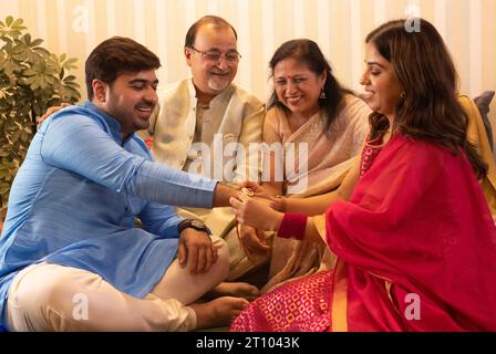 Schwester fesselt Rakhi am Handgelenk ihres Bruders an Raksha Bandhan, während die Eltern nebenan sitzen Stockfoto