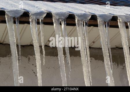 Scharfe Eiszapfen und geschmolzener Schnee hängen von den Dachtrassen. Wunderschöne, transparente Eiszapfen, die langsam über ein Dach gleiten. Stockfoto
