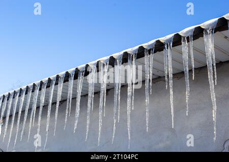 Scharfe Eiszapfen und geschmolzener Schnee hängen von den Dachtrassen. Wunderschöne, transparente Eiszapfen, die langsam über ein Dach gleiten. Stockfoto