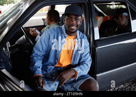 Junger, lächelnder männlicher Hip Hop-Tänzer in schwarzer Mütze, Jeansjacke und Shorts sitzt auf dem Vordersitz des Autos und blickt auf die Kamera gegen seine Freunde Stockfoto