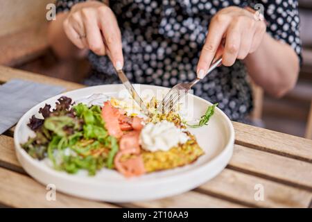 Frau schneidet Zucchini-Pfannkuchen mit Avocado, Käsesahne, Lachs und Ei. Gesundes Frühstück, Eiweiß. Restaurant-Gericht. Frühstück im Café bei Sonnenschein Stockfoto