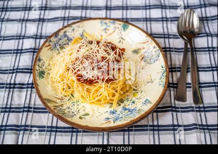 Spaghetti mit Hackfleisch, Tomatensauce (Spaghetti Bolognese) und geriebenem Käse auf Zierteller, Gabel und Löffel auf karierter Tischdecke. Stockfoto