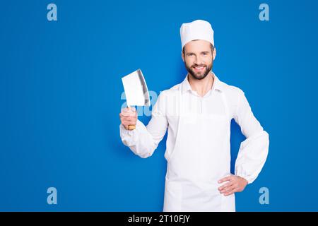 Porträt eines positiv fröhlichen, gutaussehenden Metzgers mit Hackbeil im Arm, der die Hand an der Taille hält, in die Kamera schaut, isoliert auf grauem Hintergrund Stockfoto