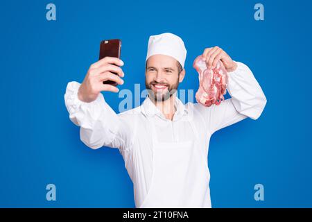 Porträt eines attraktiven, gutaussehenden Metzgers mit Bartstoppel-Selfie auf der Frontkamera des Smartphones, mit frischem Fleisch in der Hand, Blogger mit Video Stockfoto