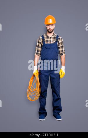 Vollkörperporträt in voller Größe, ein hübscher, strenger Elektriker mit Borsten im Overall, Hemd, gewalztes Seil in der Hand halten, in die Kamera schauen Stockfoto